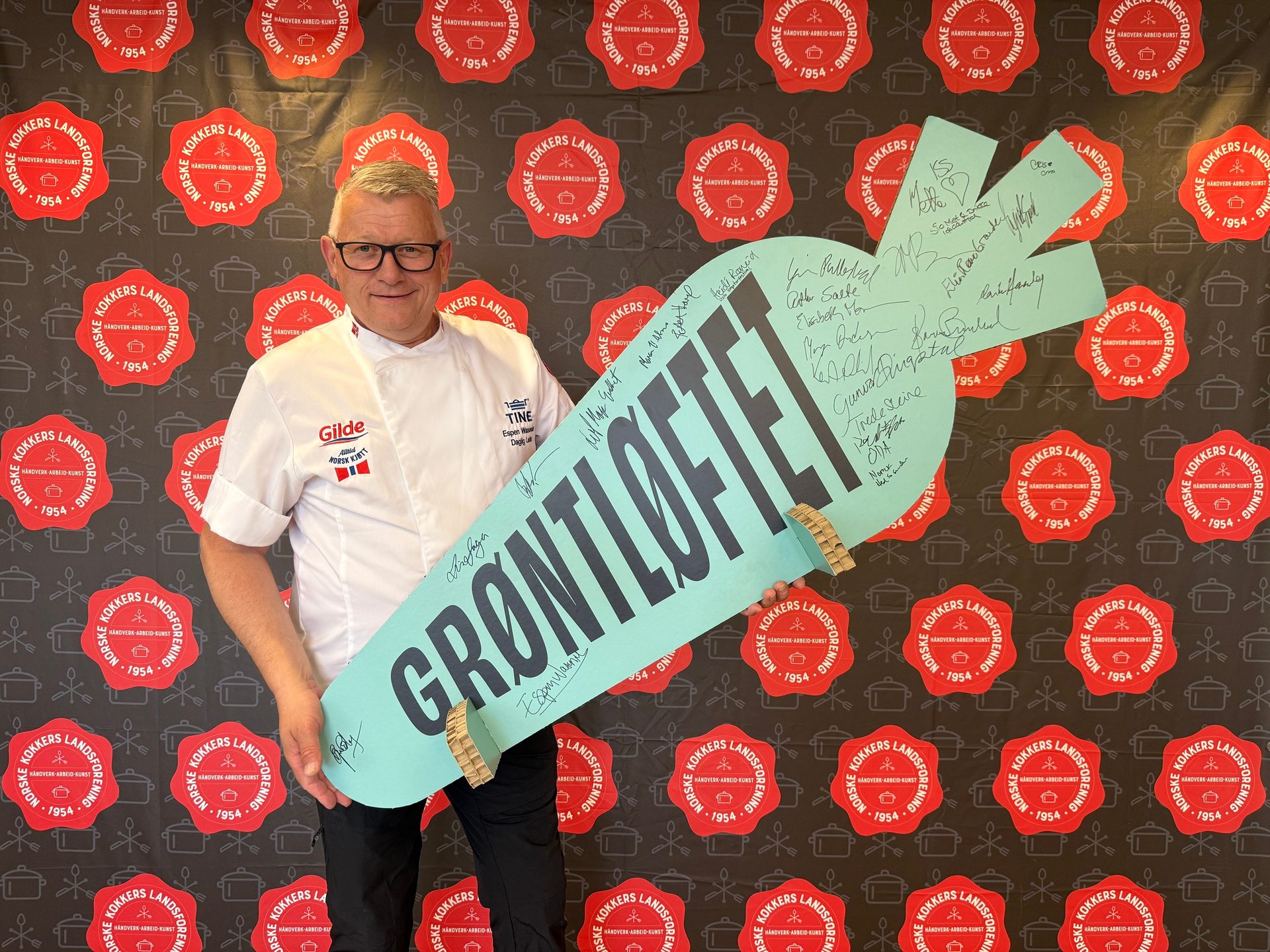 A chef in white uniform holds a large, carrot-shaped sign with 'GRØNTLOFTET' text and signatures, against a patterned backdrop.