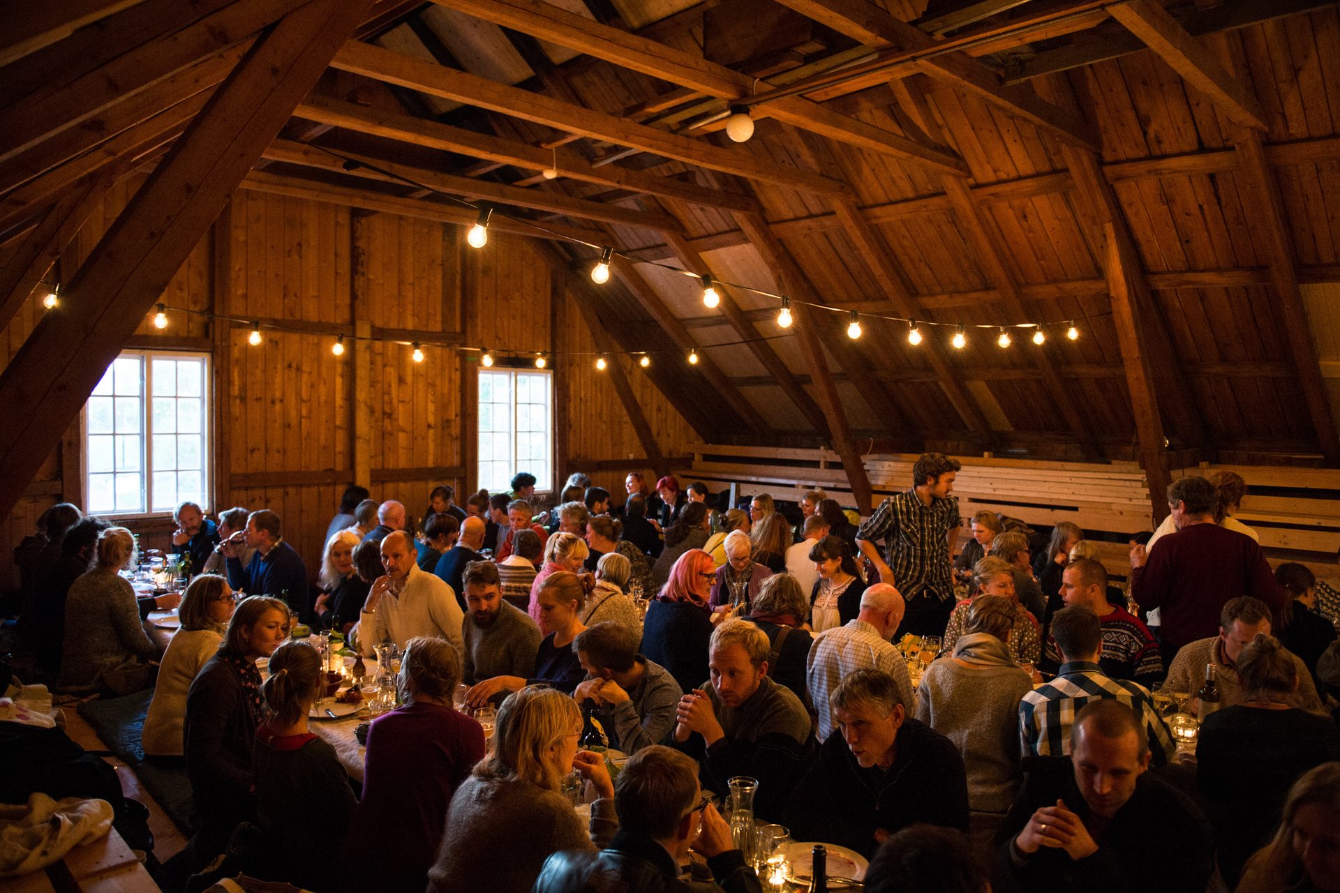 A large group of people dine and socialize in a warm, rustic wooden hall illuminated by string lights.