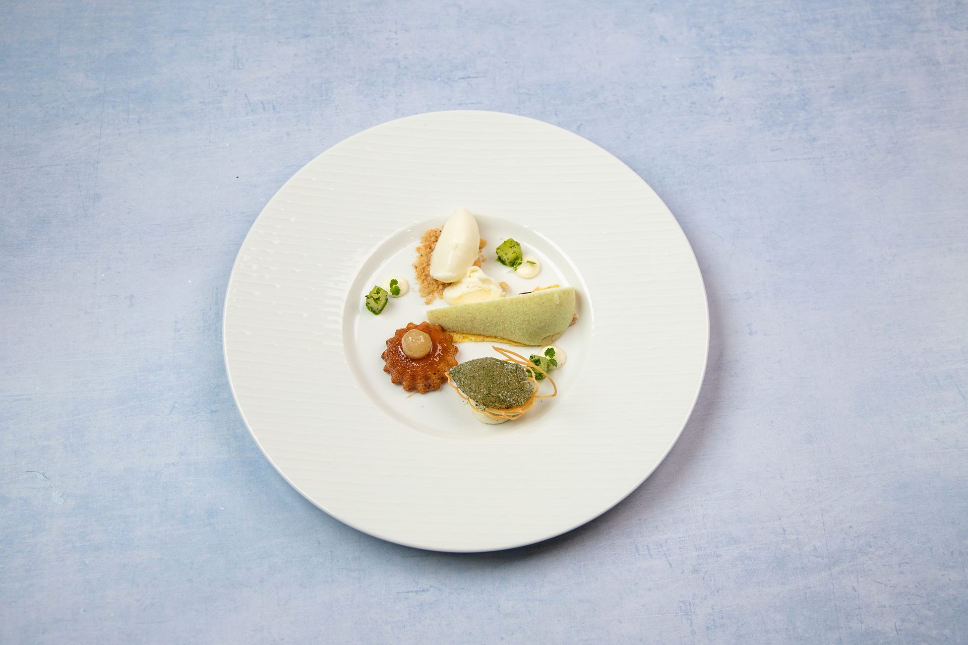 A white plate with an artfully arranged green, white, and golden dessert, garnished with crumbs.