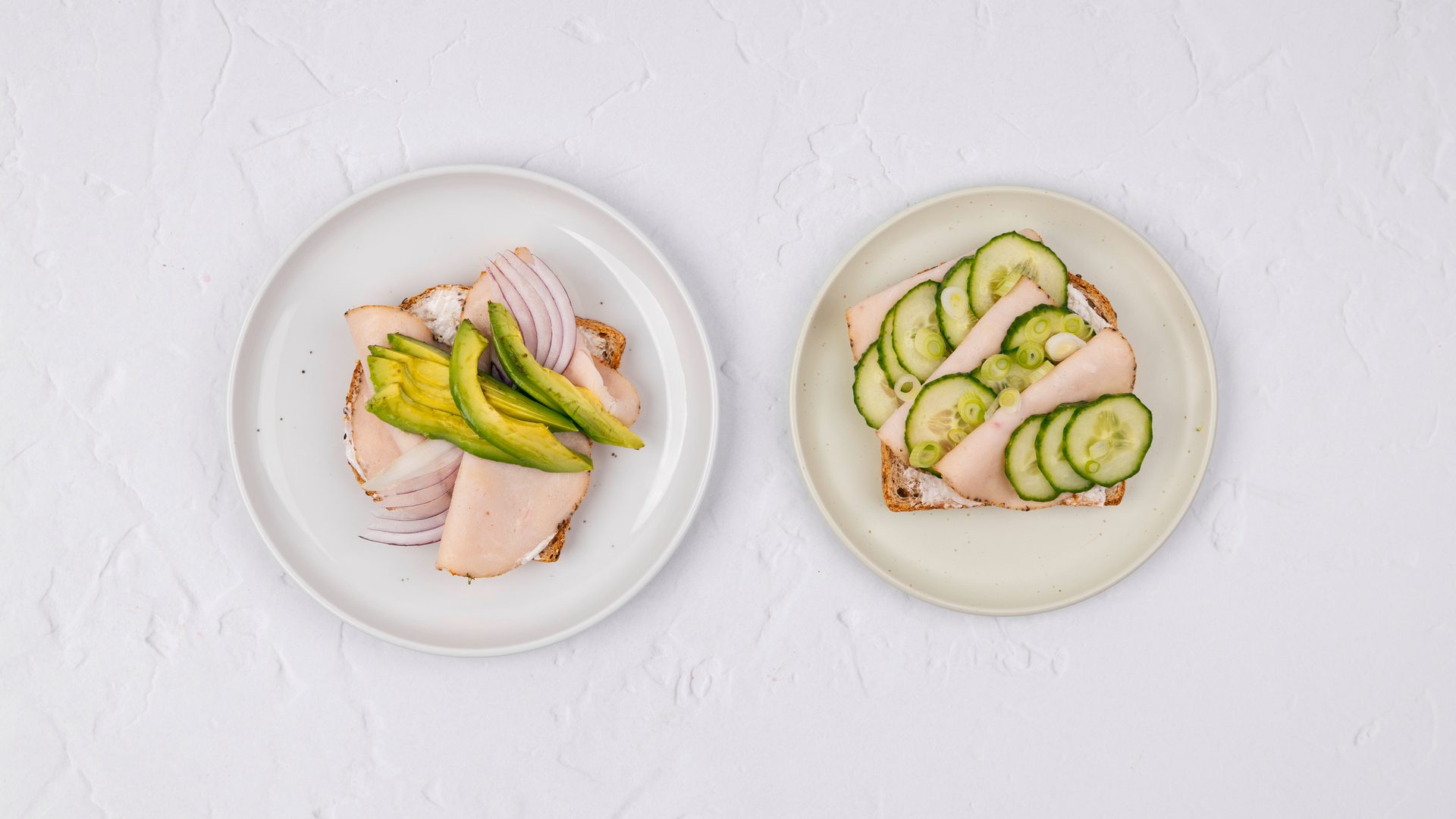 Two open-faced turkey sandwiches, one with avocado and red onion, the other with cucumber and green onion.