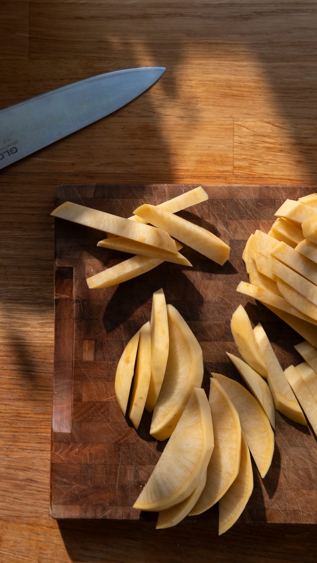 A knife and sliced root vegetables, possibly parsnips, on a wooden cutting board in sunlight.