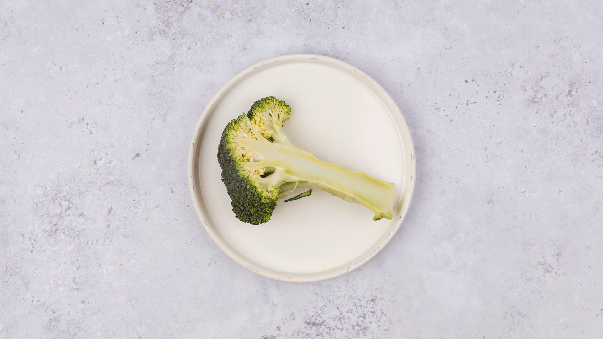 A cross-section of a broccoli floret and stem on a white plate against a grey background.