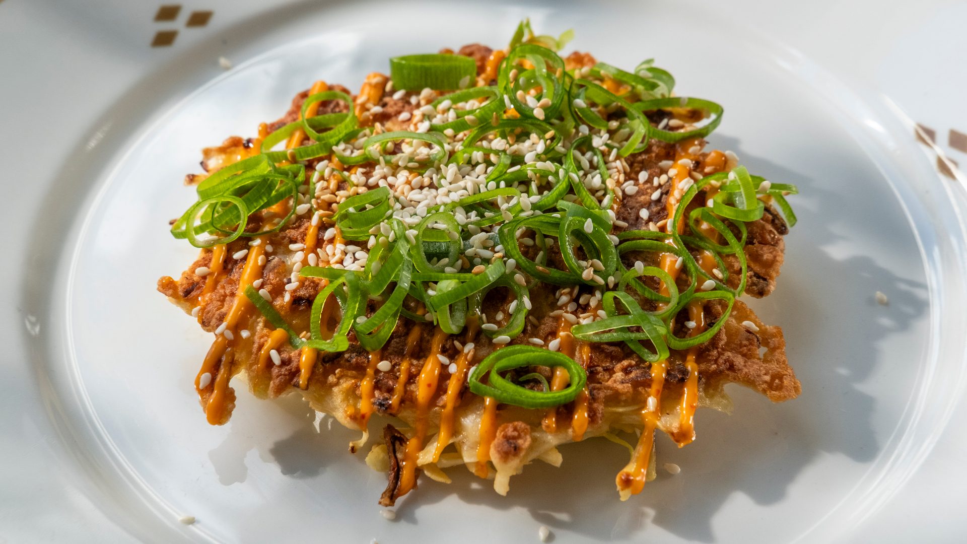 A savory pancake-style dish topped with chopped green onions, sesame seeds, and a spicy orange sauce.